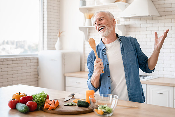 Cheerful old senior man grandfather singing and using spoon as a microphone while cooking food.